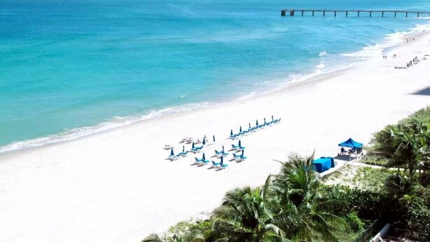 A beach with white sand, a pier extending into the ocean, and rows of blue umbrellas. - 
Miami CONDO Family-Friendly Beach Condo w/ Full Kitchen, Aventura Mall (Sunny Isles)