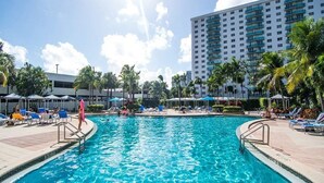A swimming pool with lounge chairs and umbrellas, surrounded by palm trees and a tall hotel building. - 
Miami CONDO Family-Friendly Beach Condo w/ Full Kitchen, Aventura Mall (Sunny Isles)