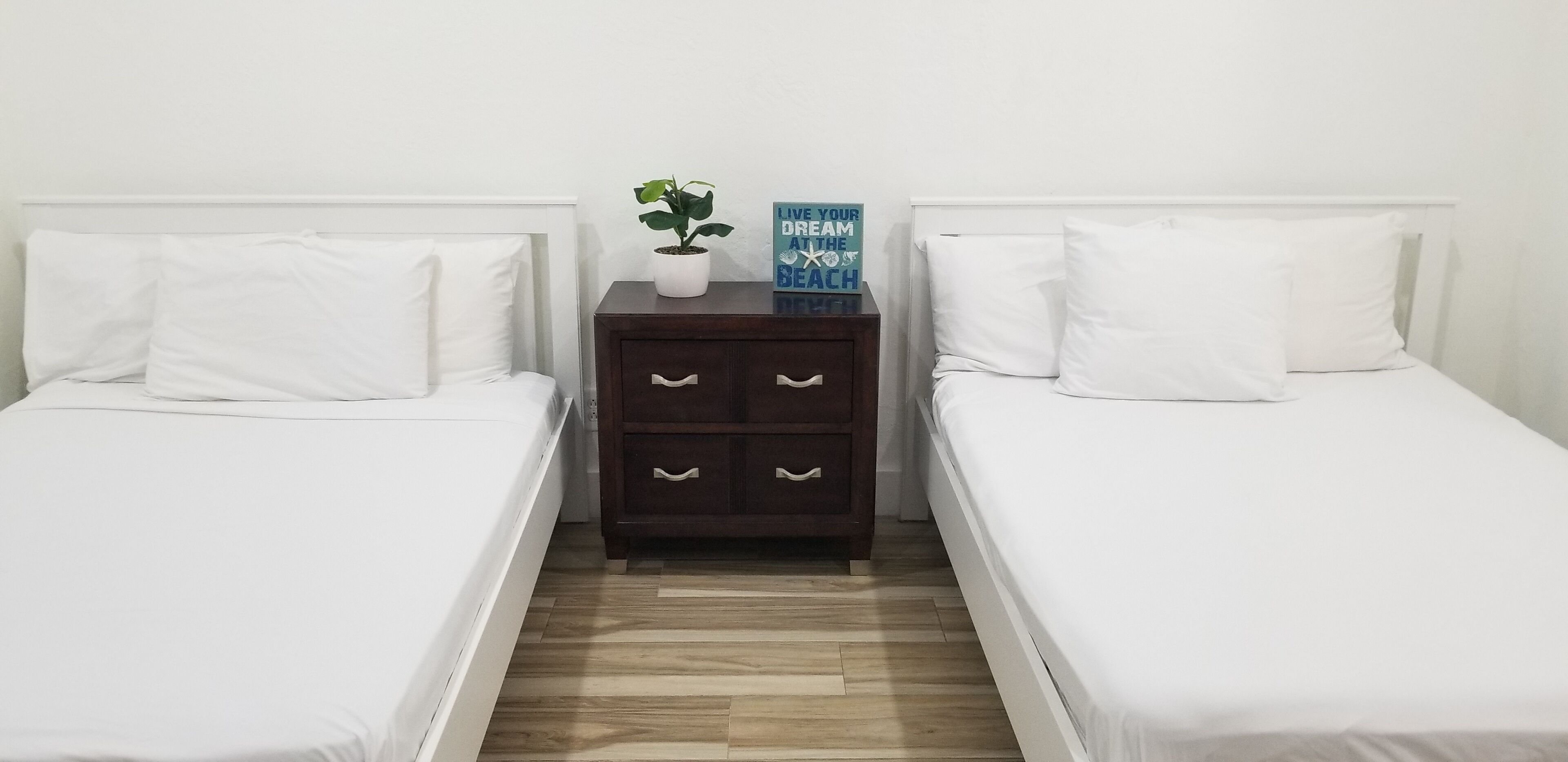 A twin bedroom with two single beds, a wooden nightstand with drawers, a potted plant, and a motivational sign.