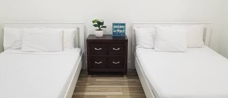 A twin bedroom with two single beds, a wooden nightstand with drawers, a potted plant, and a motivational sign.