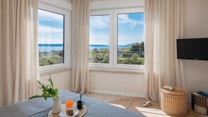 A bedroom with a large window offering a scenic view, a bed, a bedside table with a tray of breakfast, and a TV on the wall.