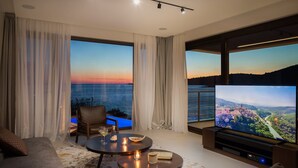 Ett modernt vardagsrum med en stor TV, bekväma sittplatser, ett litet bord med ljus och utsikt över havet vid solnedgången.