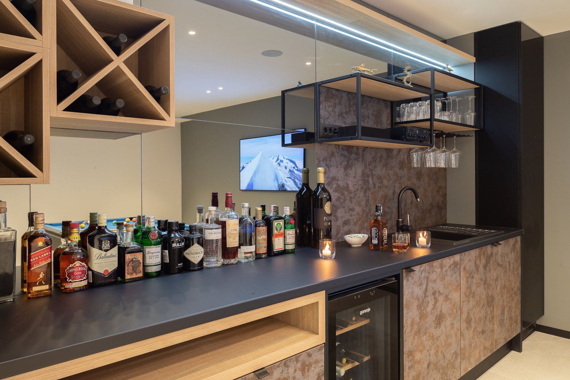 A modern bar area with a variety of liquor bottles, a wine glass rack, and a television in the background.