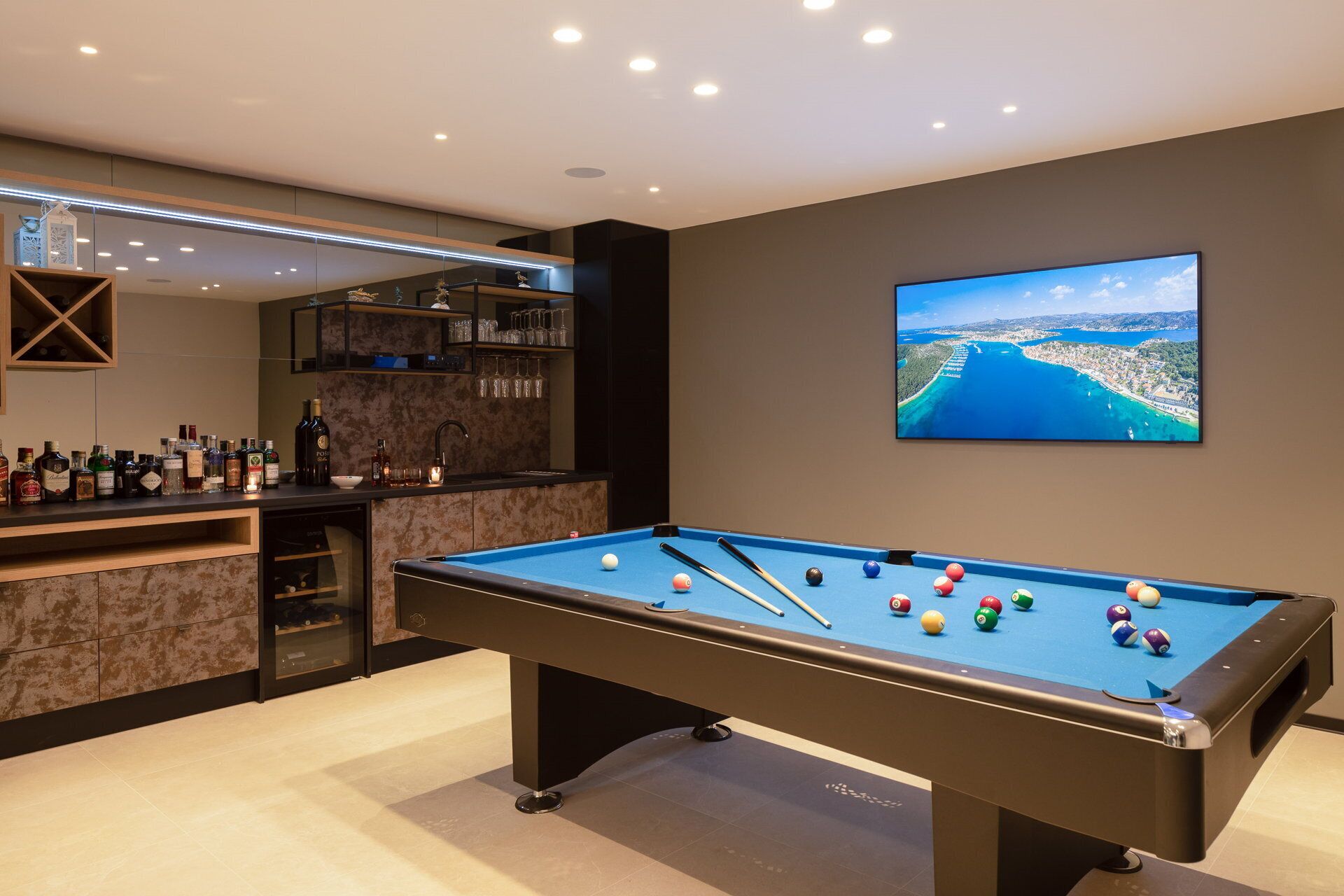 Una mesa de billar con bolas, una zona de bar con botellas y un televisor mostrando una vista panorámica.
