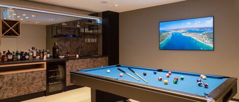 Una mesa de billar con bolas, una zona de bar con botellas y un televisor mostrando una vista panorámica.