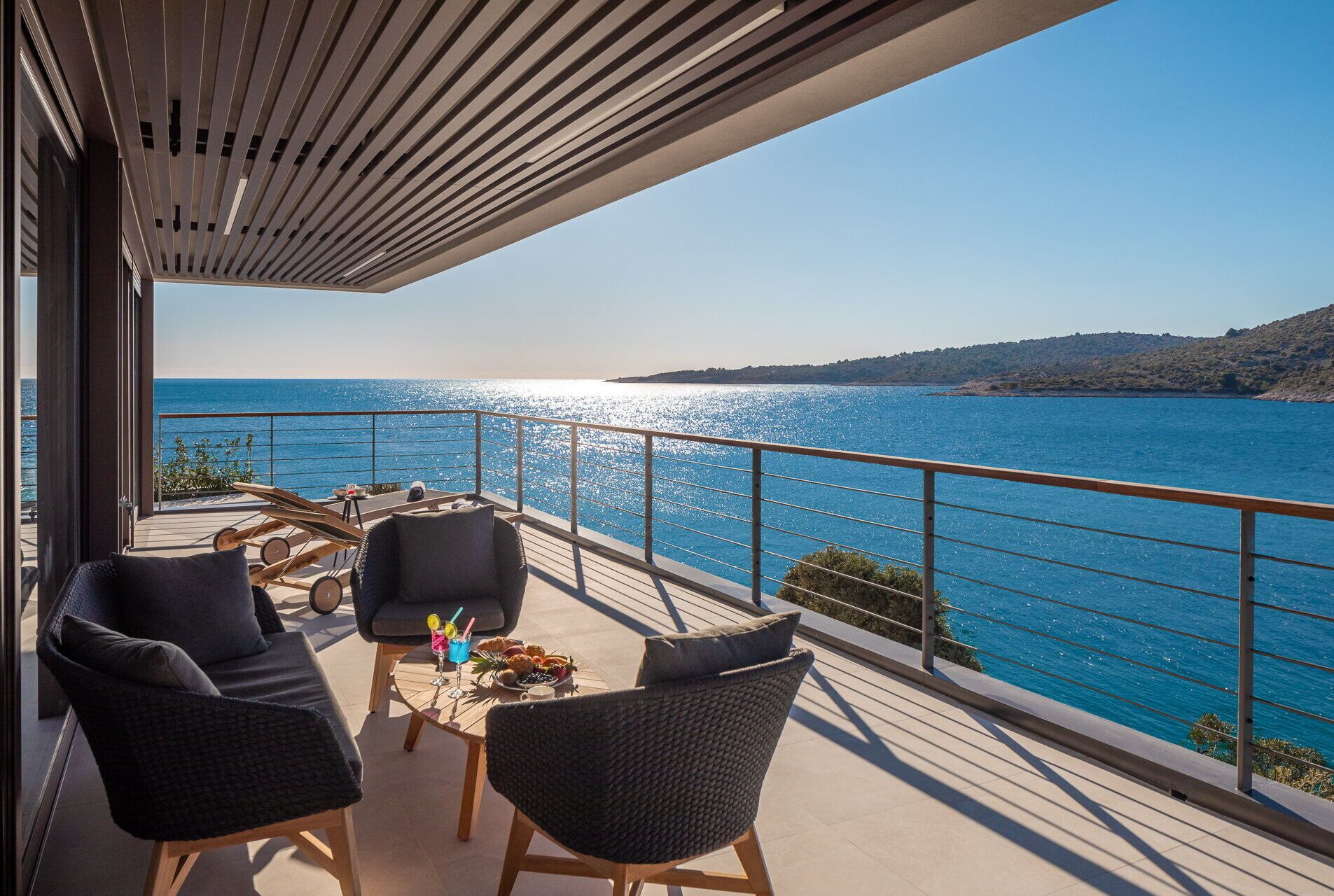Un balcon avec vue sur la mer, aménagé avec des chaises et une table où sont disposées des boissons et des collations.