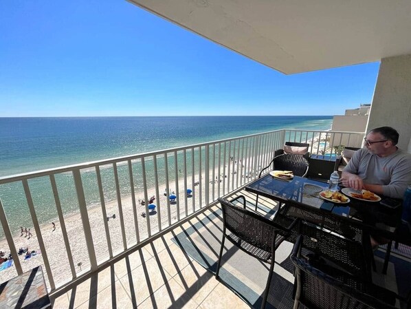 A balcony with a view of the beach and ocean, a table with plates of food, and a person sitting at the table.