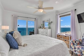 A bedroom with a bed, a dresser, a chair, a TV, and a balcony with a view of the sea.