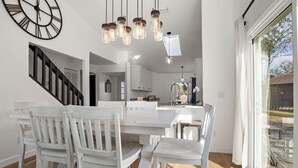 A modern kitchen with white cabinetry, a dining area with white chairs, and a large window with a view of the outdoors. - Beautiful summer open concept house that feels like home away from home, exceptionally clean with a great outdoor space and fire pit! Near speedway, airport, and beach! (Port Orange)