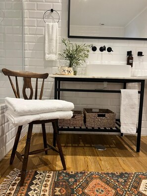 Une salle de bain avec une chaise en bois, un lavabo blanc, un miroir, une plante en pot et des paniers en osier sur une étagère.