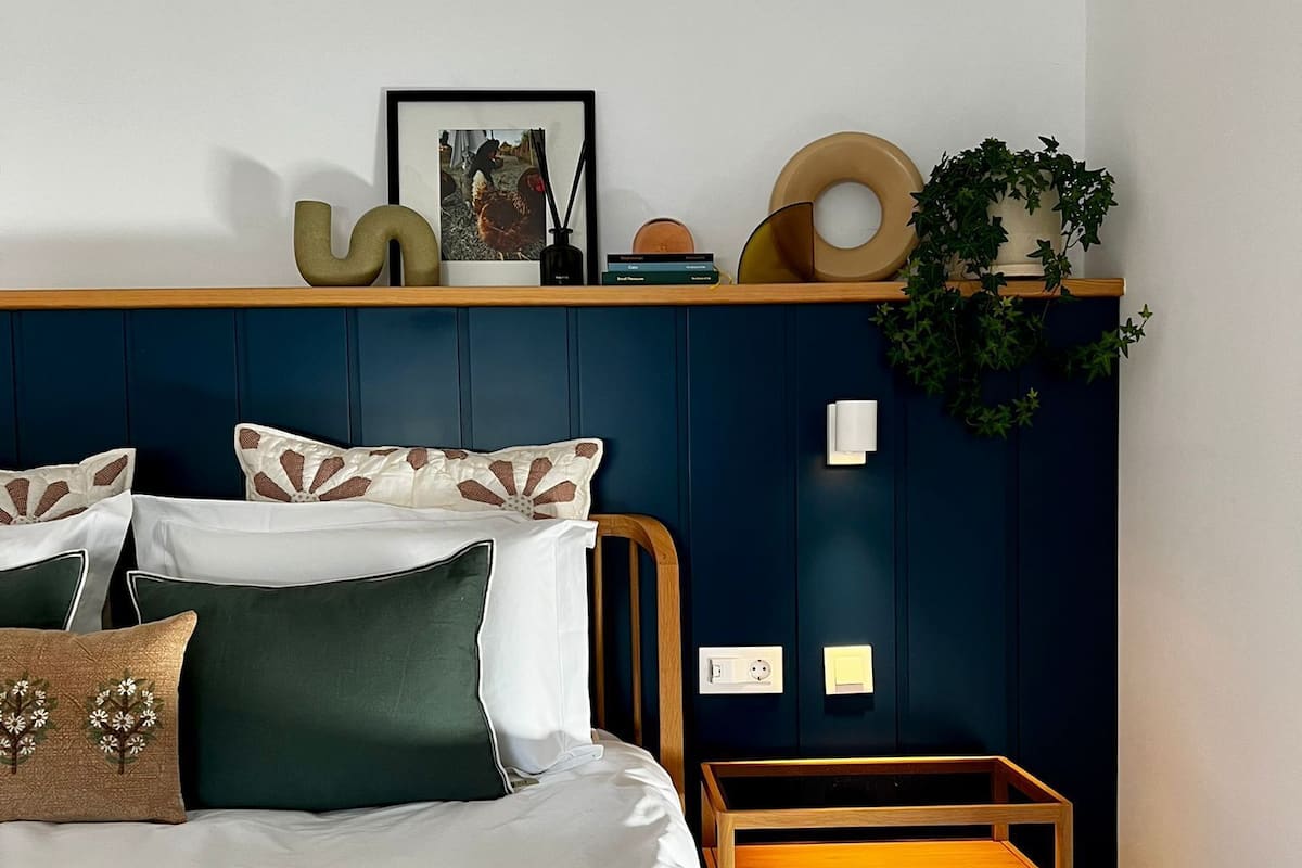 A bedroom with a bed, a bedside table, a wall-mounted shelf with decorative items, and a potted plant.