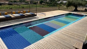 A swimming pool with a wooden deck and lounge area, surrounded by a hilly landscape.