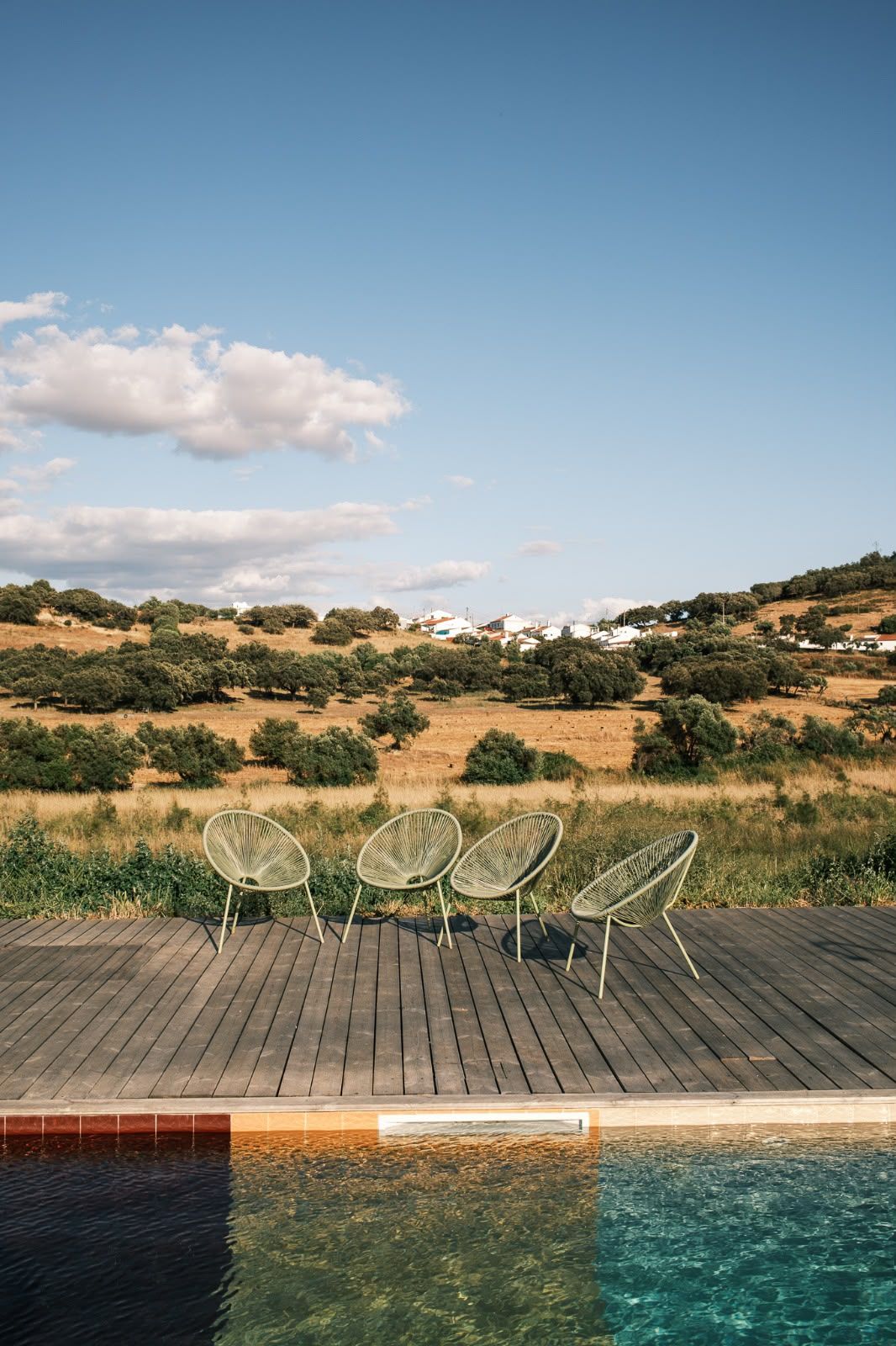 Een houten terras met drie rieten stoelen met uitzicht op een zwembad en een landschap met bomen en gebouwen.