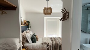 A narrow hallway leading to a bedroom with a bed, bedside tables, a hanging light fixture, and a door to a bathroom.