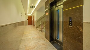 A hotel corridor with a marble floor, an elevator, and a staircase with a handrail. - Hotel Arch Suites (New Delhi)