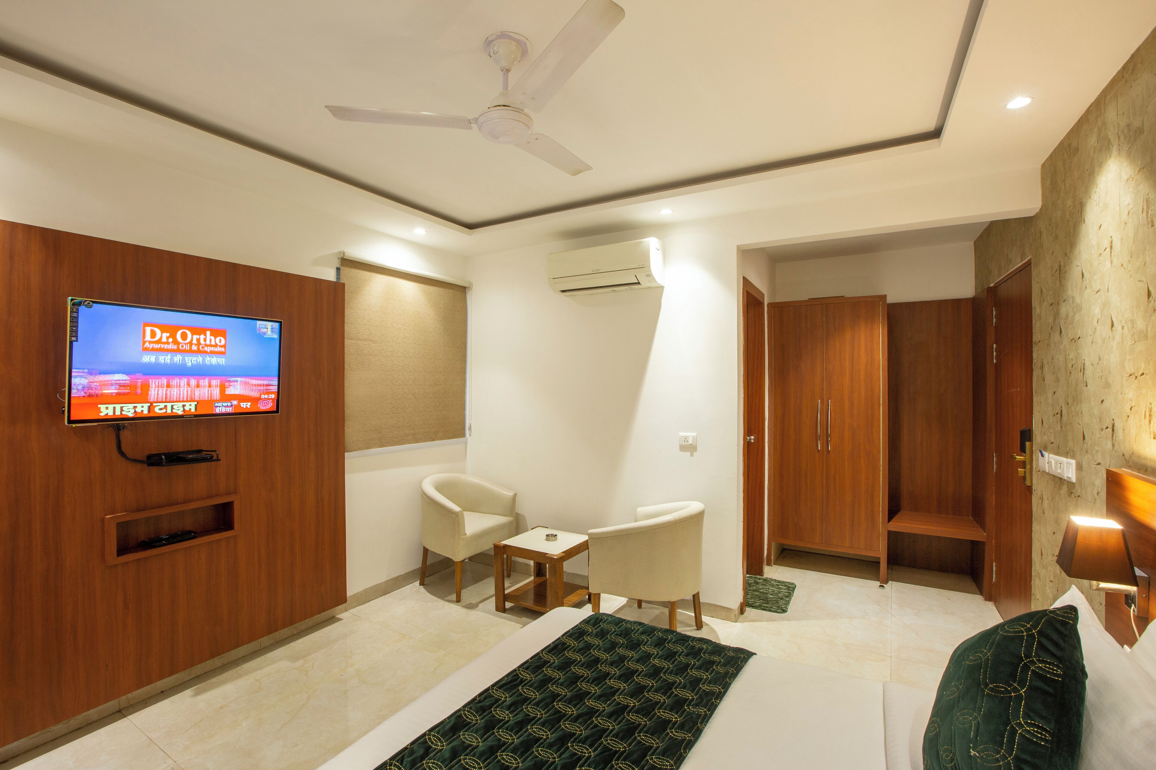 A hotel room with a bed, two chairs, a TV mounted on the wall, a ceiling fan, and a wardrobe.