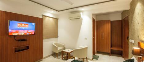 A hotel room with a bed, two chairs, a TV mounted on the wall, a ceiling fan, and a wardrobe.