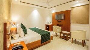 A hotel room with a bed, bedside table, chair, TV, and a small table. - Hotel Arch Suites (New Delhi)