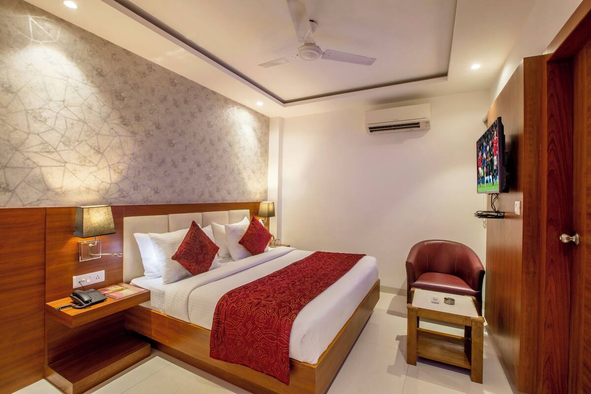 A hotel room with a wooden headboard, a bed with white linens and red accents, a small wooden nightstand with a phone and lamp, a red chair, and a ceiling fan.
