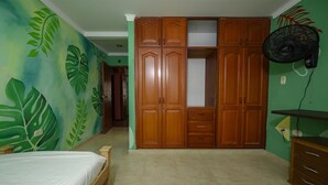 A bedroom with a bed, a wardrobe, a fan, and a green leaf mural on the wall. - Macondo Hostel (Santa Marta)
