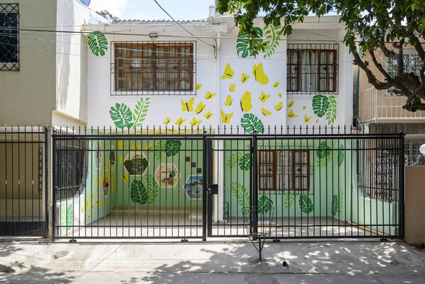 Un edificio con un mural verde y amarillo, una entrada protegida por una reja y un techo de tejas.