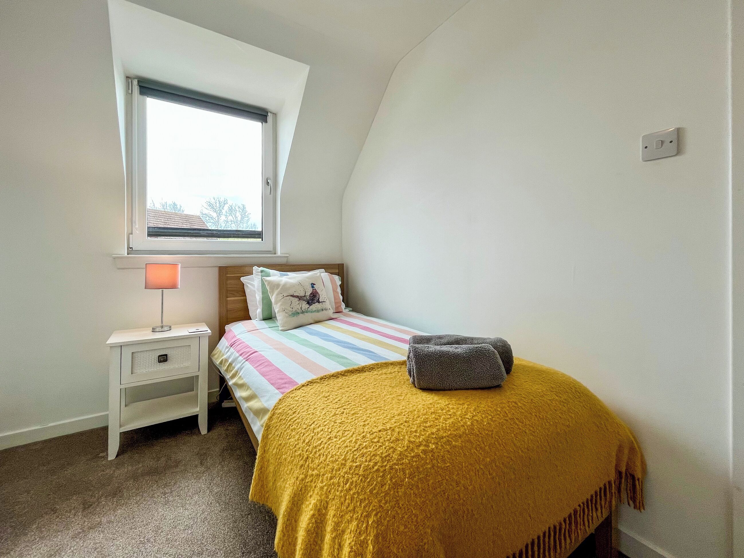 Un dormitorio con cama, mesita de noche, lámpara, ventana y toma de corriente.