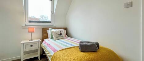 A bedroom with a bed, a nightstand, a lamp, a window, and a wall socket.