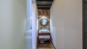 A narrow hallway leading to a bathroom with a rustic brick wall, a round mirror, a sink, and shelves with towels and shoes. - Cozy apartment in the Plaza de Callao (Madrid)