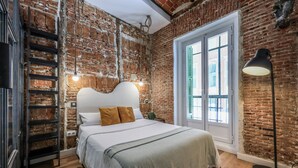 A bedroom with a brick wall, a bed with white bedding, a green throw, a black floor lamp, and a window with green shutters. - Cozy apartment in the Plaza de Callao (Madrid)