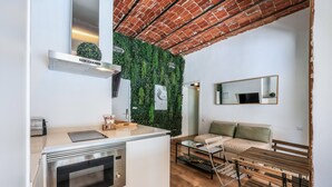 A compact kitchen with a built-in oven, a small countertop, and a wall-mounted exhaust hood. There is a wooden bench and a glass table in the living area, and a green wall with a small plant. - Cozy apartment in the Plaza de Callao (Madrid)