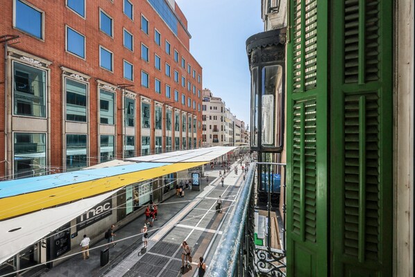 Una estación de tranvía con un tranvía moderno, gente caminando y varios edificios, incluyendo un edificio de ladrillo rojo y una estructura verde con contraventanas.