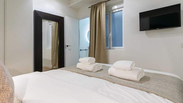 Een hotelkamer met een bed, twee opgerolde handdoeken, een aan de muur bevestigde televisie en een raam met gordijnen.