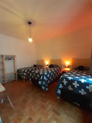 A small, cozy room with two beds, each with a cross-patterned bedspread, a wooden floor, a single hanging light fixture, and a small table with a chair. - Quarto Retrô (São Paulo)