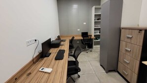 A modern office space with a long wooden desk, multiple chairs, and a computer setup. There is a whiteboard, a shelf with storage boxes, and a cabinet with drawers. - Quarto Retrô (São Paulo)