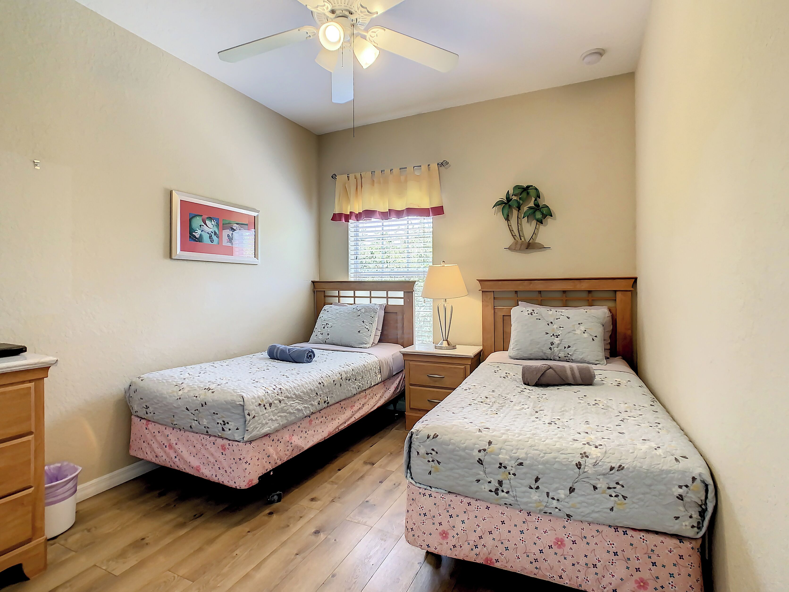 A bedroom with two single beds, a ceiling fan, a window with curtains, a bedside lamp, and a wall-mounted plant.