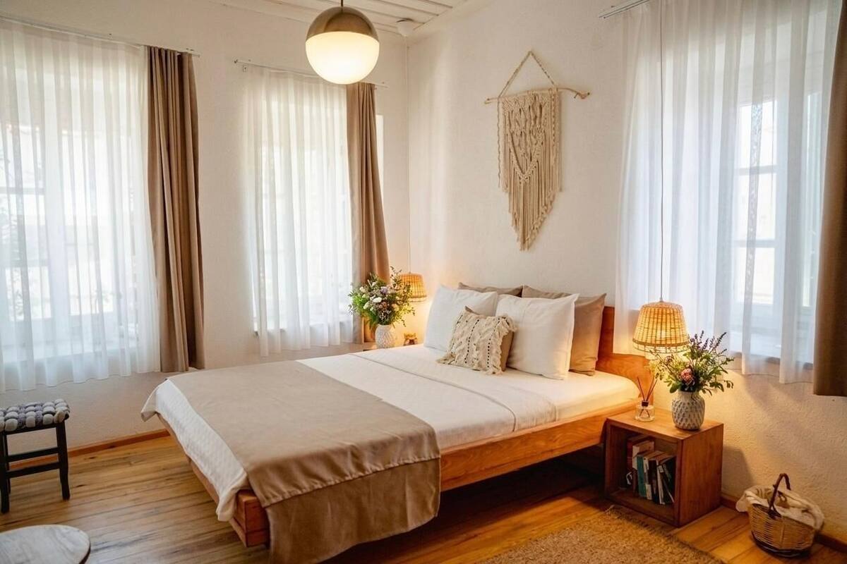 Une chambre à coucher avec un lit, des tables de chevet, une décoration murale en macramé et une fenêtre avec des rideaux.