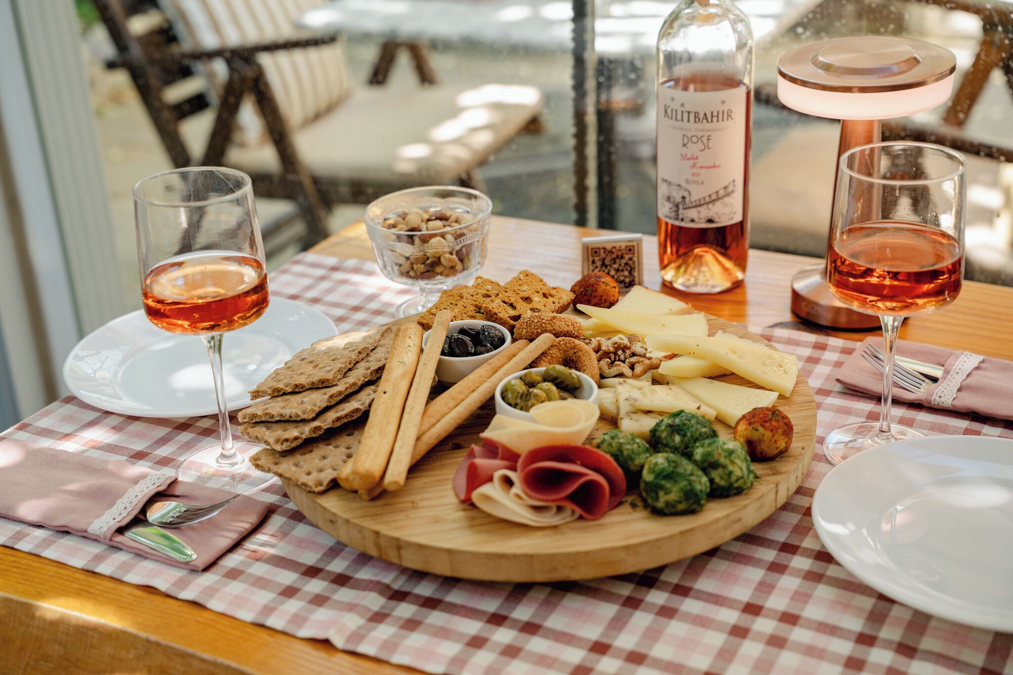 Una mesa con una variedad de quesos, carnes y galletas, acompañada de copas de vino rosado, sobre un mantel a cuadros.
