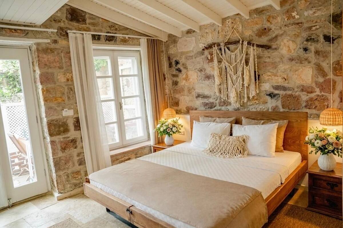 Une chambre avec un mur en pierre, un lit avec une literie blanche, une tête de lit en bois et une décoration murale en macramé.