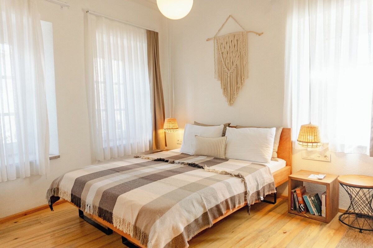 Une chambre à coucher avec un lit, des lampes de chevet, une décoration murale en macramé et une petite table d’appoint en bois sur laquelle sont posés des livres.