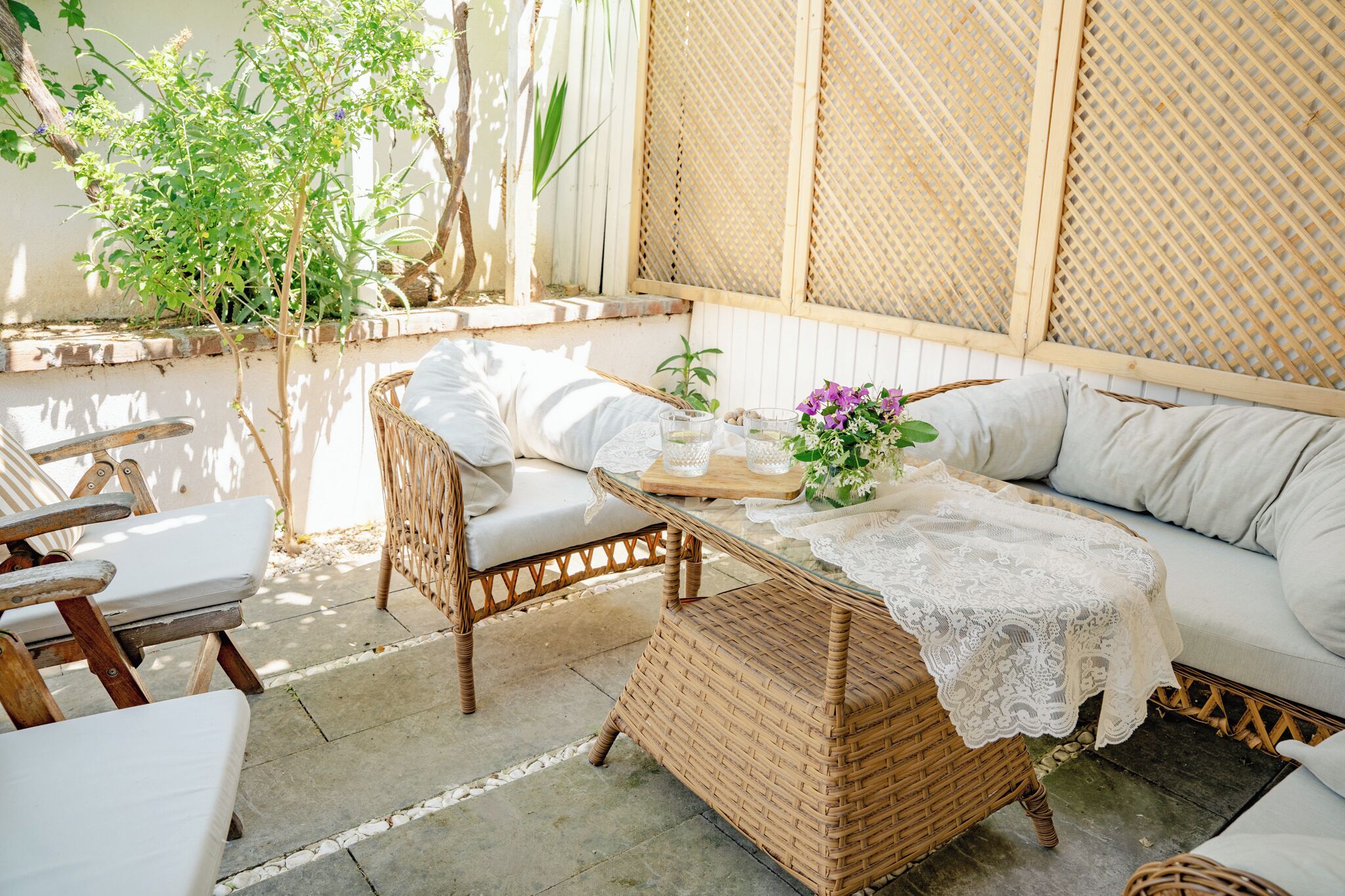 Un canapé en osier avec des coussins blancs, une table avec un plateau en verre et une nappe en dentelle, et une chaise en osier.