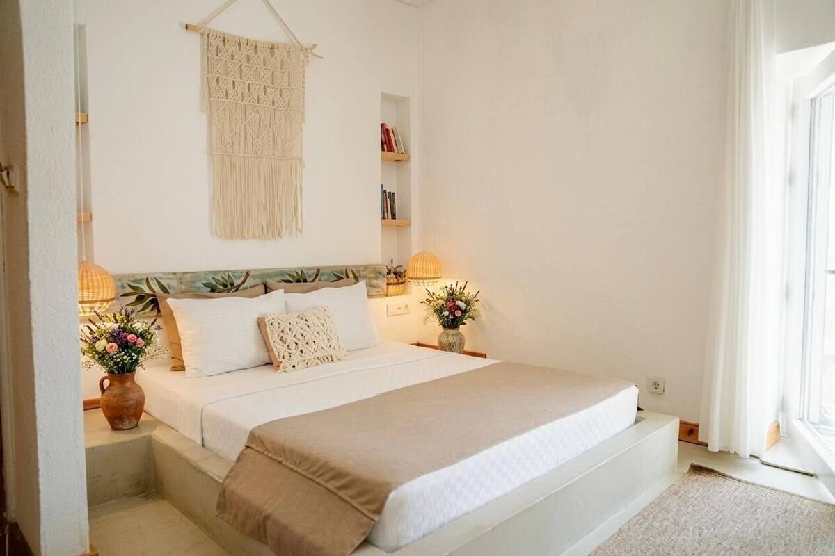 Une chambre à coucher avec un lit, des lampes de chevet, une décoration murale en macramé, une bibliothèque et une fenêtre avec des rideaux.