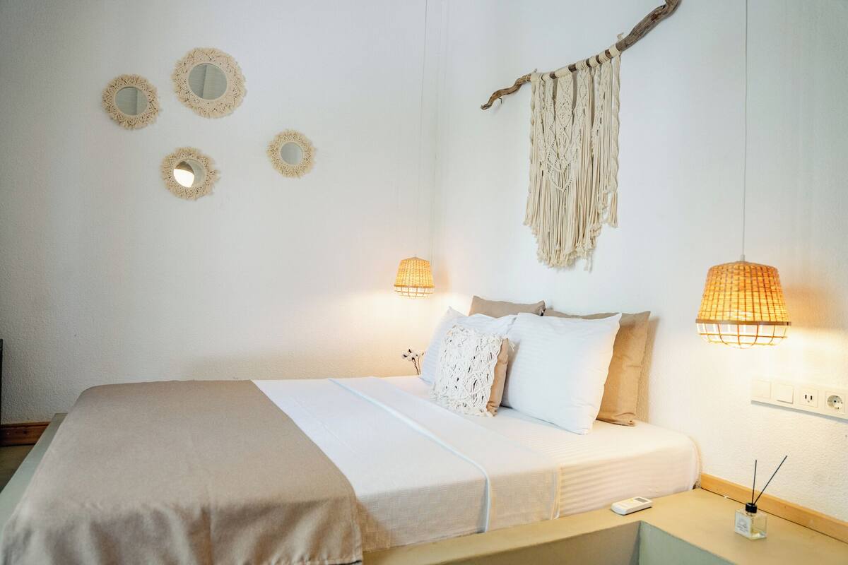 Un lit bien fait, recouvert d’une couvre-lit blanche et beige, avec deux lampes de chevet et une décoration murale en macramé.