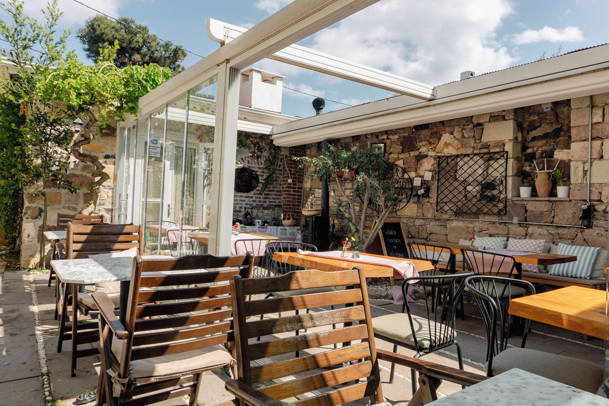 Un café al aire libre con mesas y sillas de madera, paredes de piedra y un cerramiento de cristal.