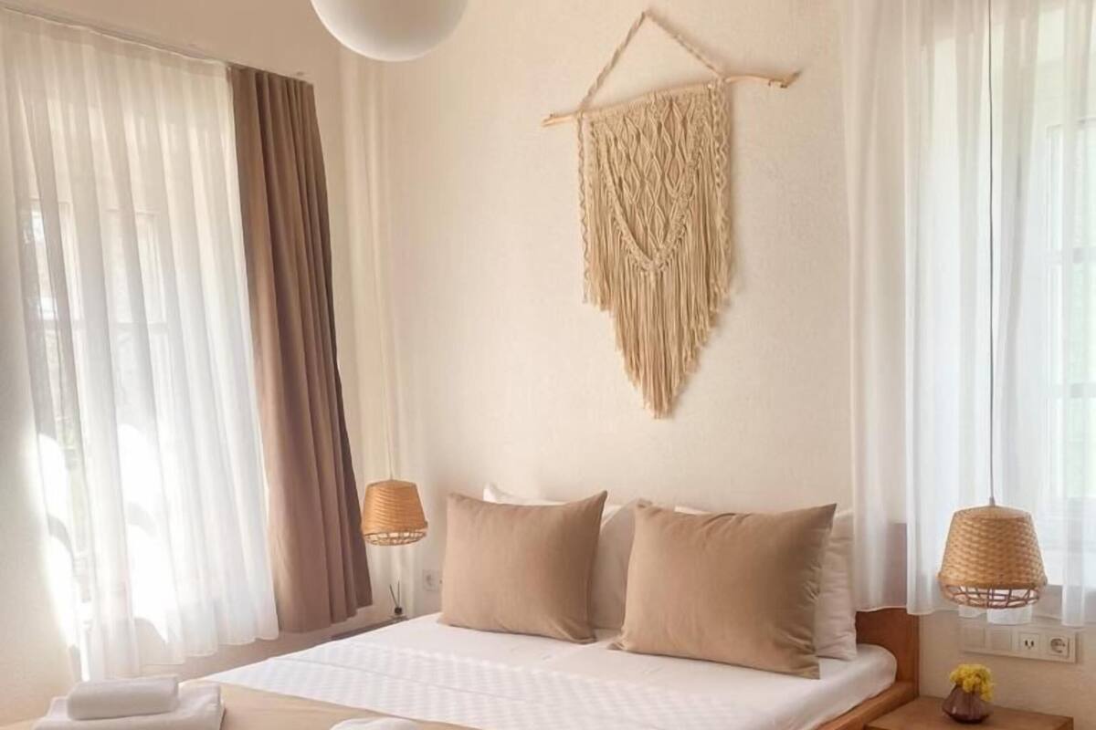 Un lit bien fait, avec des oreillers, une lampe de chevet, une décoration murale en macramé et une petite table de chevet en bois sur laquelle sont posés des livres.