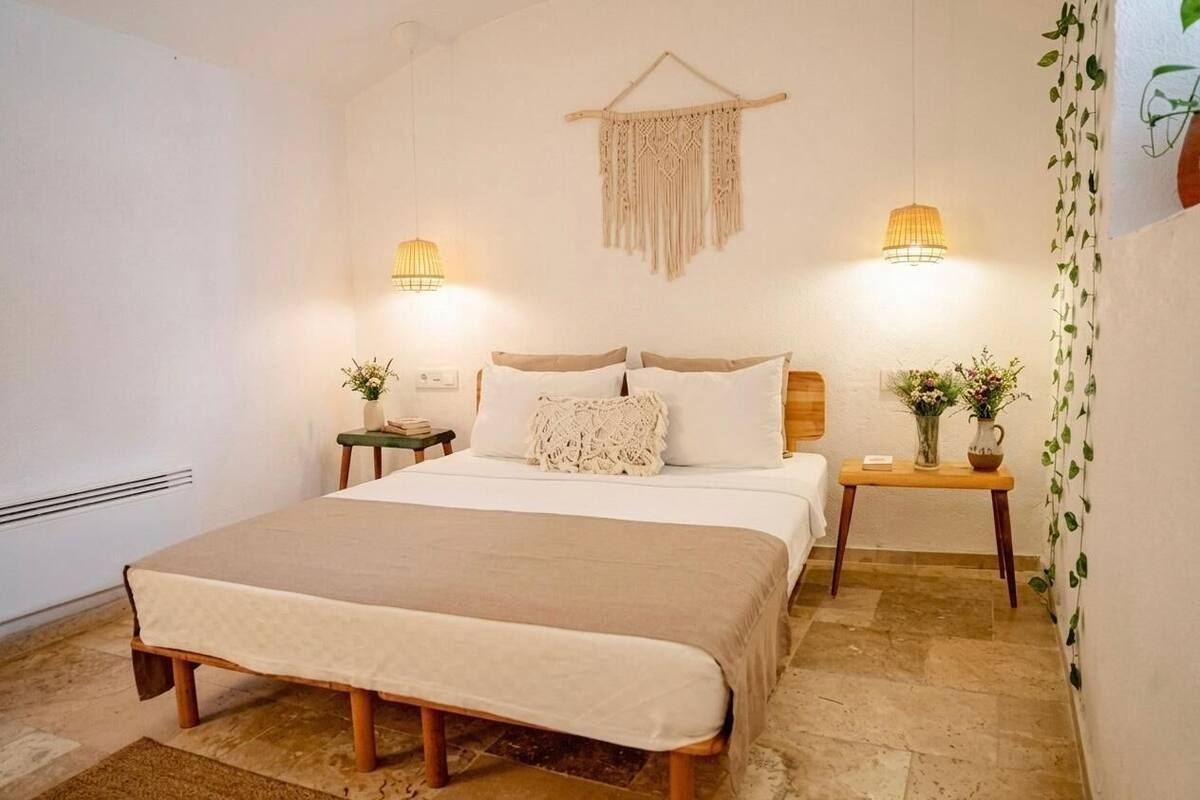 Une chambre à coucher avec un lit, des tables de chevet, des macramés accrochés au mur et des plantes suspendues.