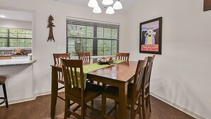 A dining area with a wooden table, chairs, a chandelier, and a window with blinds. - Retreat To Cardinals Cabin (Bryan)