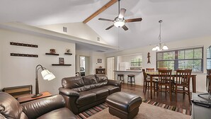 A spacious living room with a leather sofa, a dining area with wooden chairs, a ceiling fan, and a kitchen in the background. - Retreat To Cardinals Cabin (Bryan)