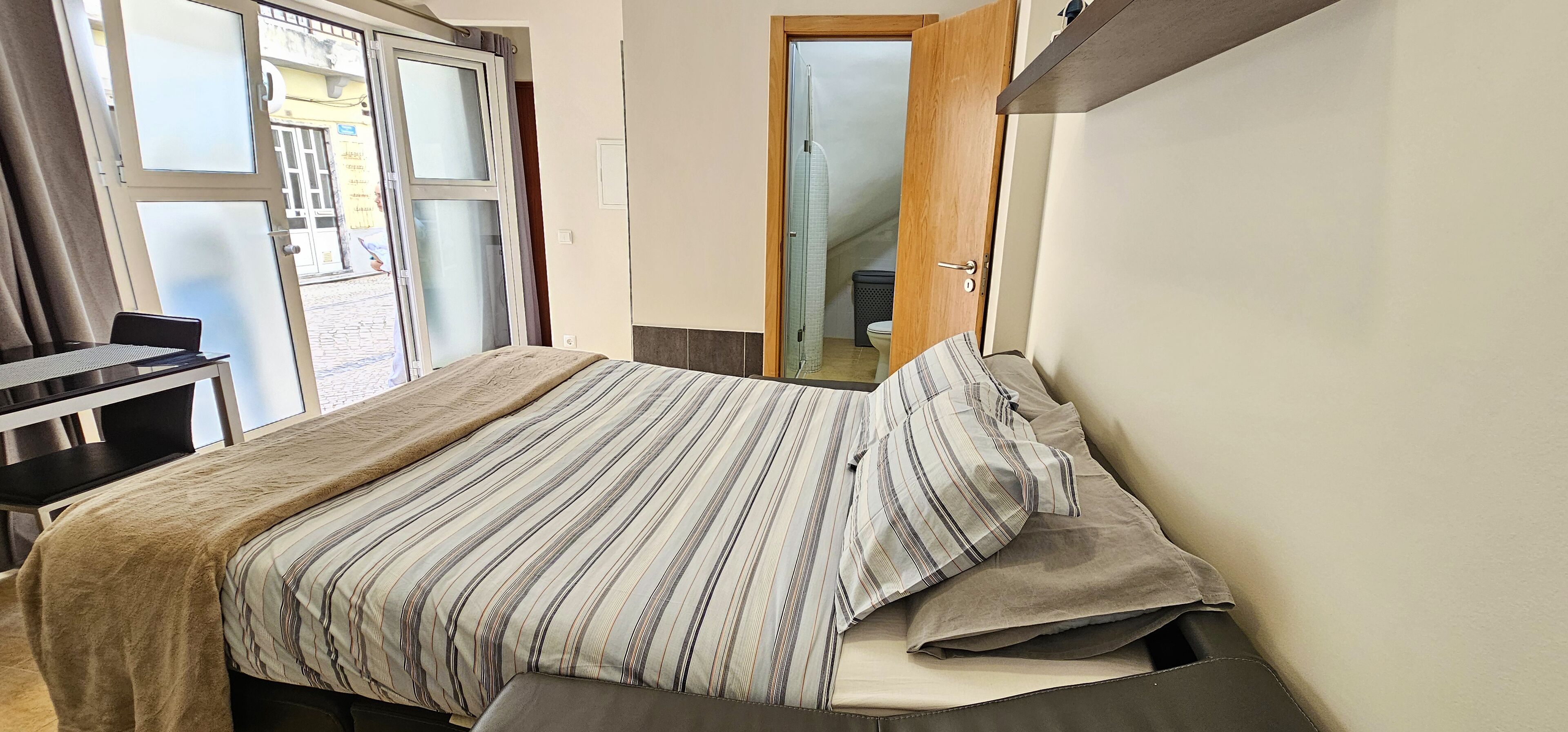 A small, neatly arranged bedroom with a bed, a bedside table, and a chair. A window with curtains is visible, and a door leads to a bathroom.