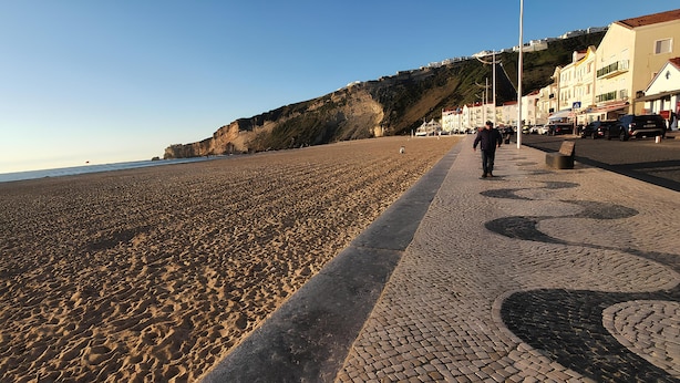 A beach with a paved walkway, buildings in the background, and a clear sky. -  Sunao (Nazaré)