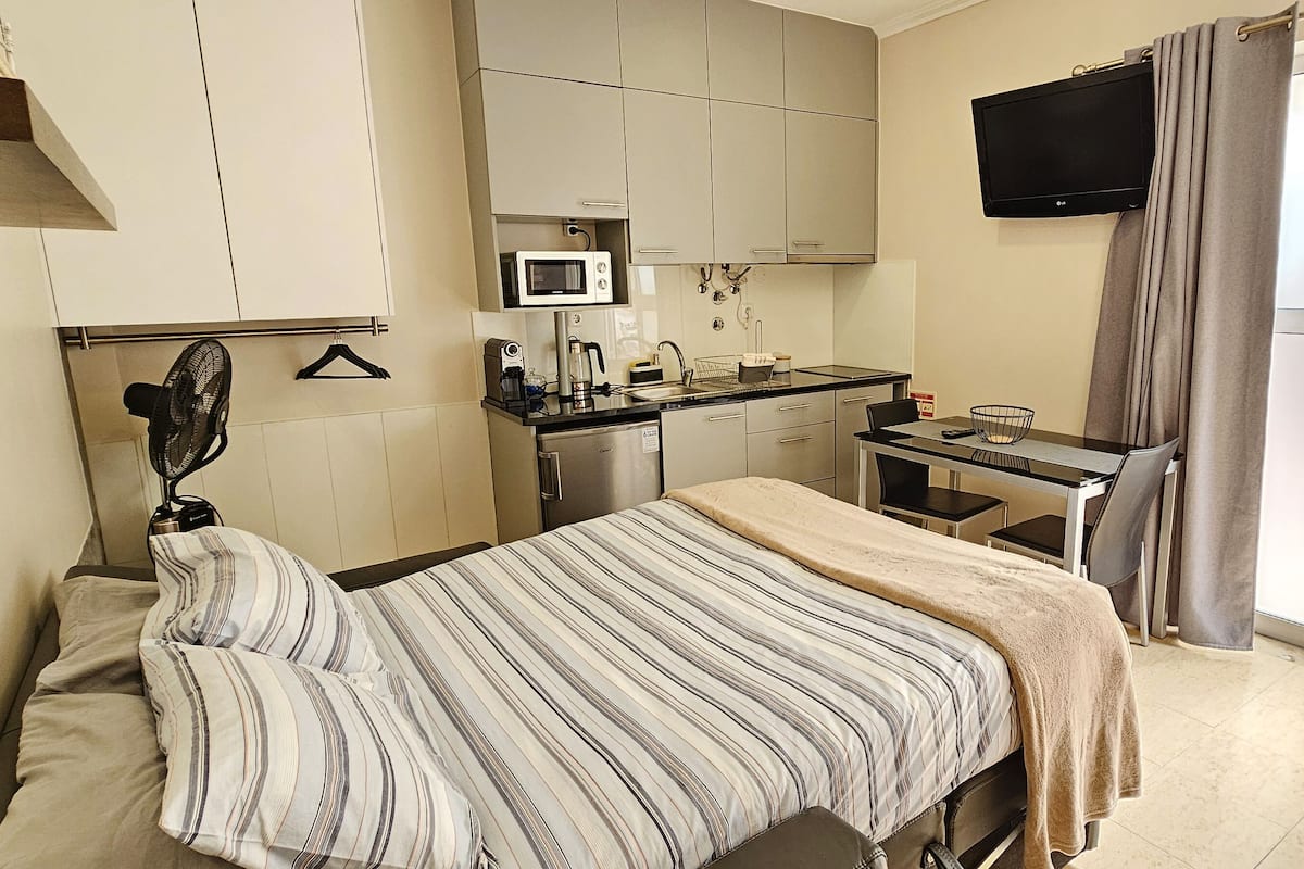 A compact hotel room with a bed, a desk, a kitchenette, a TV, and a sliding glass door to the outside.
